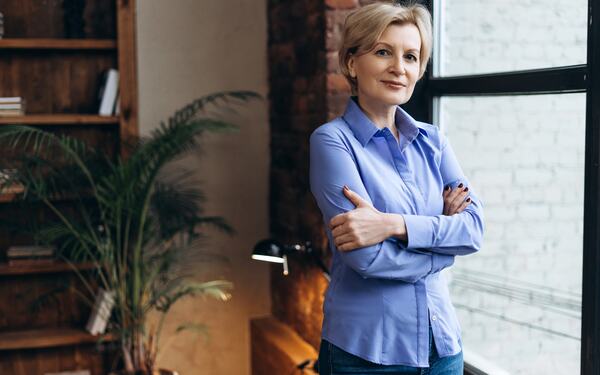 A woman in a blue shirt, standing thoughtfully — calm professional presence.