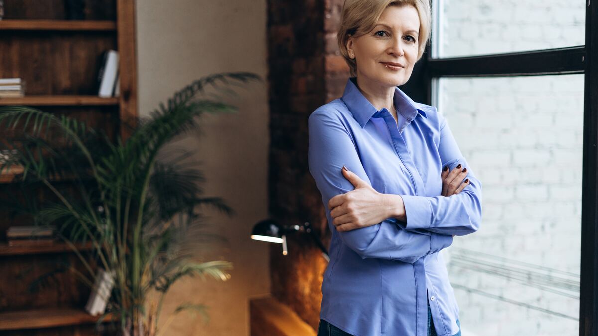 A woman in a blue shirt, standing thoughtfully — calm professional presence.